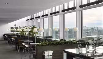 Modern cafeteria with large windows and city views. Tables and chairs are aligned near windows, surrounded by plants. A row of lamps illuminates the area. A buffet counter with covered dishes is visible in the foreground.