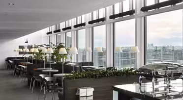 Modern cafeteria with large windows and city views. Tables and chairs are aligned near windows, surrounded by plants. A row of lamps illuminates the area. A buffet counter with covered dishes is visible in the foreground.