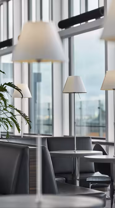 Modern lounge area with black leather seating and small round tables, lit by white table lamps. Large windows offer a view of a cityscape with a prominent tower in the distance. Green plants add a touch of nature to the space.