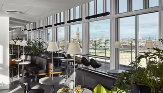 A modern dining area with large windows offers a city skyline view. The space features black and green seating, round tables with lamps, and lush indoor plants. A meal with drinks is set on a nearby table.