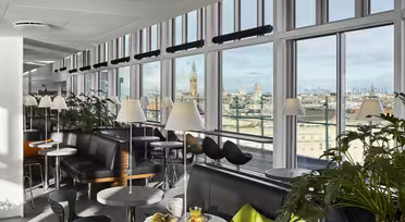 A modern dining area with large windows offers a city skyline view. The space features black and green seating, round tables with lamps, and lush indoor plants. A meal with drinks is set on a nearby table.