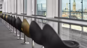 Row of modern black and golden chairs facing large windows with a cityscape view. A historic clock tower is visible through the glass, and the outside is cloudy. The setting appears to be a high-rise building, possibly an airport or office.