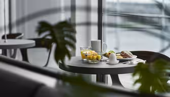 A modern dining setting with a round table near a large window. On the table are a glass of water, a bowl of fruit, a cup, a croissant, and an egg. Green foliage is partially visible in the foreground.
