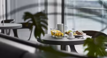 A modern dining setting with a round table near a large window. On the table are a glass of water, a bowl of fruit, a cup, a croissant, and an egg. Green foliage is partially visible in the foreground.