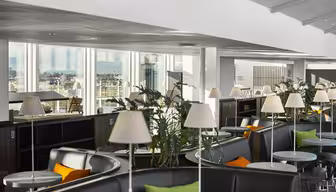 Modern lounge with curved seating, small round tables, and white lamps. Potted plants decorate the area. Large windows offer a scenic city view. Bright and spacious interior with a mix of black, orange, and green accents.