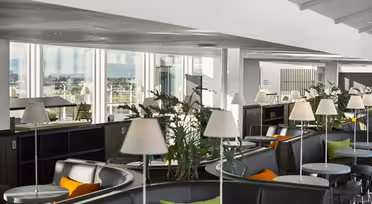 Modern lounge with curved seating, small round tables, and white lamps. Potted plants decorate the area. Large windows offer a scenic city view. Bright and spacious interior with a mix of black, orange, and green accents.