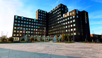 A modern, angular building with a dark facade and a pattern of square windows against a blue sky. The foreground features an open, paved area with geometric designs. Wakeup is written vertically on the building.