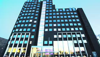 Tall modern building with a dark facade, featuring multiple square windows. The entrance is brightly lit, with glass doors and large windows. Bicycles are parked outside, and the sky is clear. The central section has a vertical line of illuminated windows.