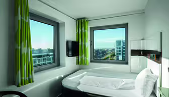 A modern hotel room with a bed, wall-mounted TV, and two large windows showing a cityscape. The room features green and white curtains and simple, sleek furnishings. Natural light illuminates the space.