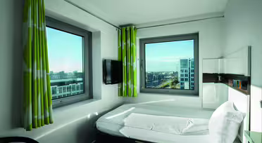 A modern hotel room with a bed, wall-mounted TV, and two large windows showing a cityscape. The room features green and white curtains and simple, sleek furnishings. Natural light illuminates the space.