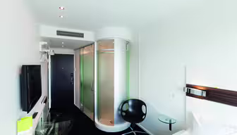 Modern hotel room with a sleek design featuring a bed, flat-screen TV, glass desk, black chair, and an enclosed circular shower. The minimalist decor includes white walls and a dark carpet, with spotlights on the ceiling.