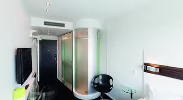 Modern hotel room with a sleek design featuring a bed, flat-screen TV, glass desk, black chair, and an enclosed circular shower. The minimalist decor includes white walls and a dark carpet, with spotlights on the ceiling.