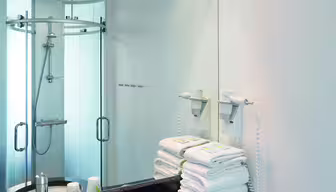 Modern white bathroom with a large mirror, a black marble countertop, and a round sink. Towels are neatly stacked beside a wall-mounted hairdryer. In the corner, theres a glass-enclosed shower. Soft lighting overhead brightens the space.