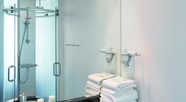 Modern white bathroom with a large mirror, a black marble countertop, and a round sink. Towels are neatly stacked beside a wall-mounted hairdryer. In the corner, theres a glass-enclosed shower. Soft lighting overhead brightens the space.