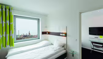 Modern hotel room with a bed next to a window showing a cityscape view. The room is bright, featuring white bedding and green-striped curtains. A small glass-topped side table is beside the bed, and a wall-mounted TV is in the adjacent room.