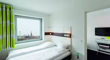 Modern hotel room with a bed next to a window showing a cityscape view. The room is bright, featuring white bedding and green-striped curtains. A small glass-topped side table is beside the bed, and a wall-mounted TV is in the adjacent room.