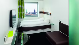 A modern hotel room with a brown sofa, wall-mounted TV, and a bed under a large window. The window offers a view of a cityscape with historic buildings. The room has white walls and minimalist decor, including a small glass table and patterned curtains.