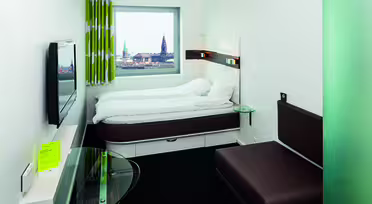 A modern hotel room with a brown sofa, wall-mounted TV, and a bed under a large window. The window offers a view of a cityscape with historic buildings. The room has white walls and minimalist decor, including a small glass table and patterned curtains.