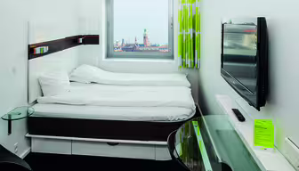 Minimalist hotel room with a double bed, wall-mounted TV, and a small glass desk. A window offers a view of a cityscape with a tall tower and distant water. The decor is modern with white bedding and lime-green curtains.