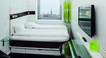 Minimalist hotel room with a double bed, wall-mounted TV, and a small glass desk. A window offers a view of a cityscape with a tall tower and distant water. The decor is modern with white bedding and lime-green curtains.