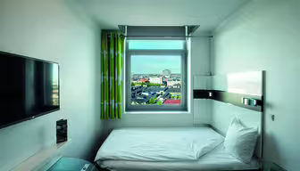 Minimalistic hotel room with a single bed placed under a window showing a cityscape. A wall-mounted TV is on the left, and the room has light green curtains. The decor is modern and simple, with a glass-topped desk partially visible.