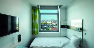 Minimalistic hotel room with a single bed placed under a window showing a cityscape. A wall-mounted TV is on the left, and the room has light green curtains. The decor is modern and simple, with a glass-topped desk partially visible.