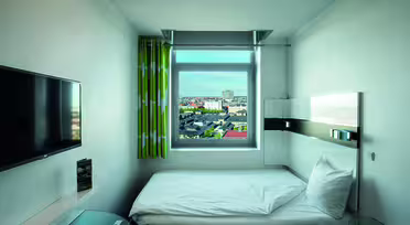 Minimalistic hotel room with a single bed placed under a window showing a cityscape. A wall-mounted TV is on the left, and the room has light green curtains. The decor is modern and simple, with a glass-topped desk partially visible.