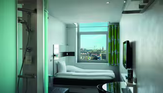 Modern hotel room with a single bed, flat-screen TV, and round glass table. A glass-enclosed shower is visible in the corner. A large window offers a cityscape view with skyscrapers and a partly cloudy sky. Bright green curtains add a pop of color.