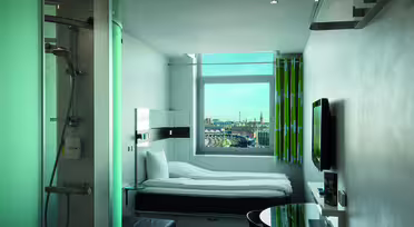Modern hotel room with a single bed, flat-screen TV, and round glass table. A glass-enclosed shower is visible in the corner. A large window offers a cityscape view with skyscrapers and a partly cloudy sky. Bright green curtains add a pop of color.