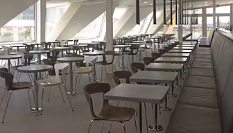 A modern cafeteria with rows of empty tables and chairs. The space features large windows allowing natural light to illuminate the area. There are long black booths on the right side and a minimalistic design throughout.
