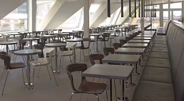 A modern cafeteria with rows of empty tables and chairs. The space features large windows allowing natural light to illuminate the area. There are long black booths on the right side and a minimalistic design throughout.