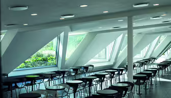 Modern cafeteria with rows of circular tables and chairs, featuring large, slanted windows allowing natural light and a view of greenery outside. The ceiling has several recessed lights.