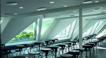 Modern cafeteria with rows of circular tables and chairs, featuring large, slanted windows allowing natural light and a view of greenery outside. The ceiling has several recessed lights.