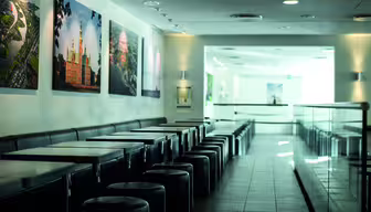 A modern, empty café with black tables and stools lined up against a wall. The wall displays large framed photos of landmarks. The floor is tiled, and soft lighting creates a calm atmosphere. A counter with a glass barrier is on the right.