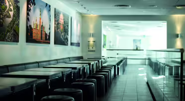 A modern, empty café with black tables and stools lined up against a wall. The wall displays large framed photos of landmarks. The floor is tiled, and soft lighting creates a calm atmosphere. A counter with a glass barrier is on the right.