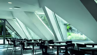 Modern cafe with sleek design, featuring angular, floor-to-ceiling windows that allow natural light to fill the space. The room includes empty round tables and dark chairs, creating an open and minimalist atmosphere.