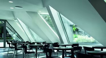 Modern cafe with sleek design, featuring angular, floor-to-ceiling windows that allow natural light to fill the space. The room includes empty round tables and dark chairs, creating an open and minimalist atmosphere.