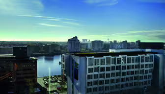 View of a cityscape featuring modern buildings by a body of water under a clear blue sky. The foreground shows a white building, with a partially visible street and parked cars below.
