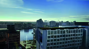 View of a cityscape featuring modern buildings by a body of water under a clear blue sky. The foreground shows a white building, with a partially visible street and parked cars below.