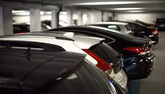 A row of parked cars in a dimly lit underground parking garage. The vehicles are lined up closely, with their fronts facing away from the camera. The environment appears spacious with white walls and a low ceiling.