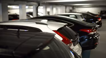 A row of parked cars in a dimly lit underground parking garage. The vehicles are lined up closely, with their fronts facing away from the camera. The environment appears spacious with white walls and a low ceiling.