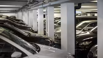 Cars parked closely in an indoor parking garage. The image shows rows of dark-colored vehicles under artificial lighting, with structural columns separating the parking spaces. The area appears to be a well-lit, organized facility.