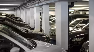 Cars parked closely in an indoor parking garage. The image shows rows of dark-colored vehicles under artificial lighting, with structural columns separating the parking spaces. The area appears to be a well-lit, organized facility.