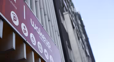 A modern building with a red sign displaying icons for parking, accessibility, reception, and café facilities. The sign includes the word Wakeup. The building exterior features vertical white and black elements. Sky visible in the background.