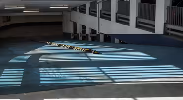 A parking garage ramp is lit by sunlight streaming through vertical slats, creating striped shadows on the teal floor. A yellow speed bump runs across the ramp, and the background shows fencing and concrete walls.