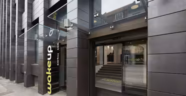 Modern hotel entrance with dark brown tiles and glass canopy. The word wake up is displayed vertically on the wall. The glass doors lead to a minimalist interior with stairs. Reflections of nearby buildings are visible on the glass.