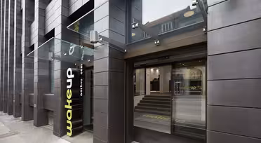 Modern hotel entrance with dark brown tiles and glass canopy. The word wake up is displayed vertically on the wall. The glass doors lead to a minimalist interior with stairs. Reflections of nearby buildings are visible on the glass.