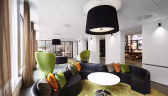 Modern lounge area with curved black sofas and vibrant green, orange, and brown cushions. Green egg-shaped chairs complement the design. Large black pendant lights hang from a white ceiling. Beige curtains and light wood flooring complete the space.