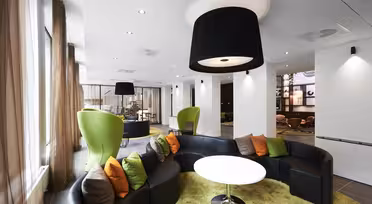 Modern lounge area with curved black sofas and vibrant green, orange, and brown cushions. Green egg-shaped chairs complement the design. Large black pendant lights hang from a white ceiling. Beige curtains and light wood flooring complete the space.
