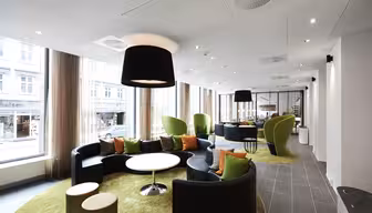 Modern lounge area with curved black sofas, green cushioned chairs, and orange pillows on green carpet. Large windows allow natural light to fill the room. Circular tables and overhead black lamps complete the cozy and stylish space.